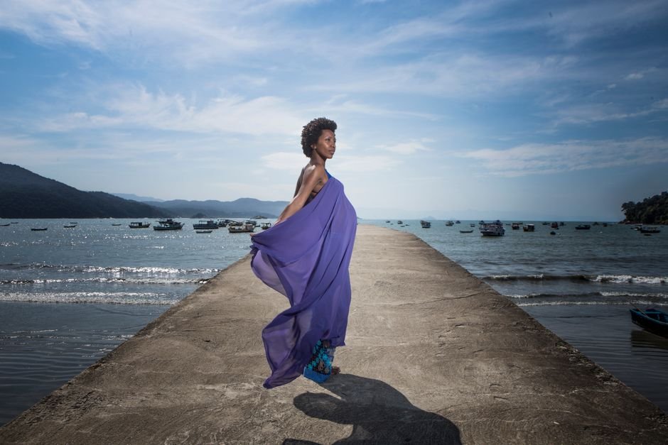 A woman gracefully poses on a pier with a flowing purple dress, evoking a sense of freedom by the ocean.