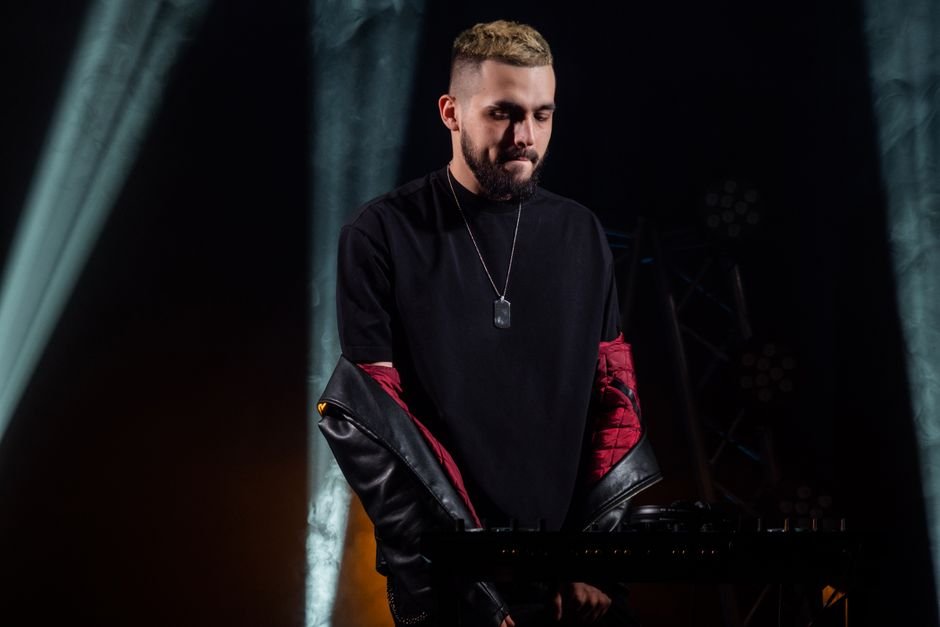 A DJ wearing a black shirt and leather jacket performing live on stage during a vibrant concert.